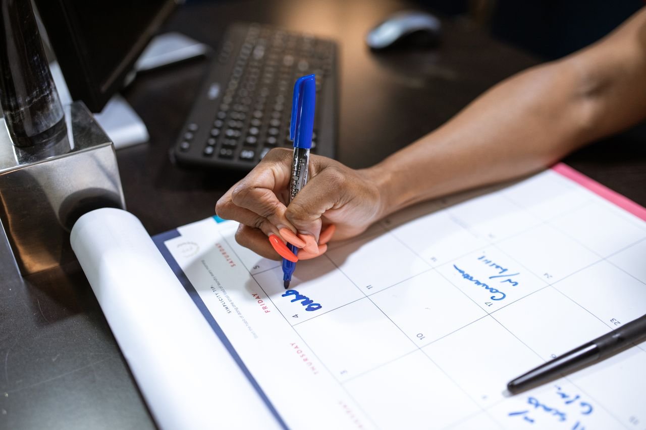 gallery-1 Detailed shot of a person's hand writing notes on a calendar using a blue marker.